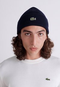 Mørkeblå strikket lue med ribbestruktur og en liten grønn krokodille-logo. Bæres med en hvit sweatshirt med en matchende logo.