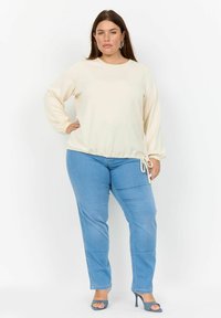 Krämfärgad långärmad topp med elstisk dragsko nederkant, kombinerad med ljusblå tighta jeans och matchande högklackade skor.