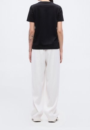 Persoon staat met de rug naar de kijker, draagt een zwart T-shirt met korte mouwen, losse witte broek en witte schoenen tegen een effen witte achtergrond.