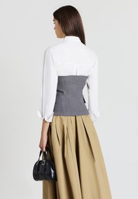 Una donna di spalle indossa una camicia bianca con corsetto grigio, una gonna plissettata beige, e tiene in mano una piccola borsa nera nella mano destra, su uno sfondo bianco.