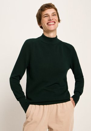 Lächelnde Person in einem dunkelgrünen Langarmpullover und beigen Hosen, die mit den Händen in den Taschen vor einem schlichten Hintergrund steht.