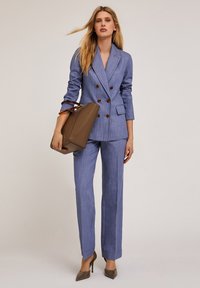 Lichtblauw dubbelgeknoopt kostuum met een op maat gemaakte pasvorm, voorzien van bruine knopen en bijpassende brede broek. Houdt een bruine handtas met accenten vast.