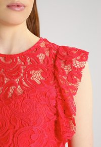 Top en dentelle rouge avec des motifs floraux complexes, un col rond et des manches volantes. Texture douce et éléments transparents visibles.