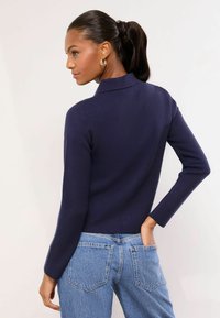 Tengerészkék kötött pulóver magas nyakkal és hosszú ujjakkal, világoskék farmernadrággal, hátsó zsebekkel. Lágy textúra látható.