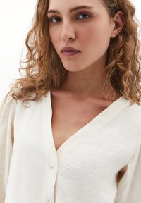 Licht crème blouse met een diepe V-hals, knoopsluiting en textuurstof. Zachte, vloeiende mouwen en een ontspannen silhouette.