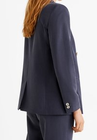 Merihenkinen bleiseri rakenteellisella istuvuudella, jossa on yksi nappi, viistetyt läpät ja nappihihnat. Sujuva kangas ja vähäinen yksityiskohtaisuus.