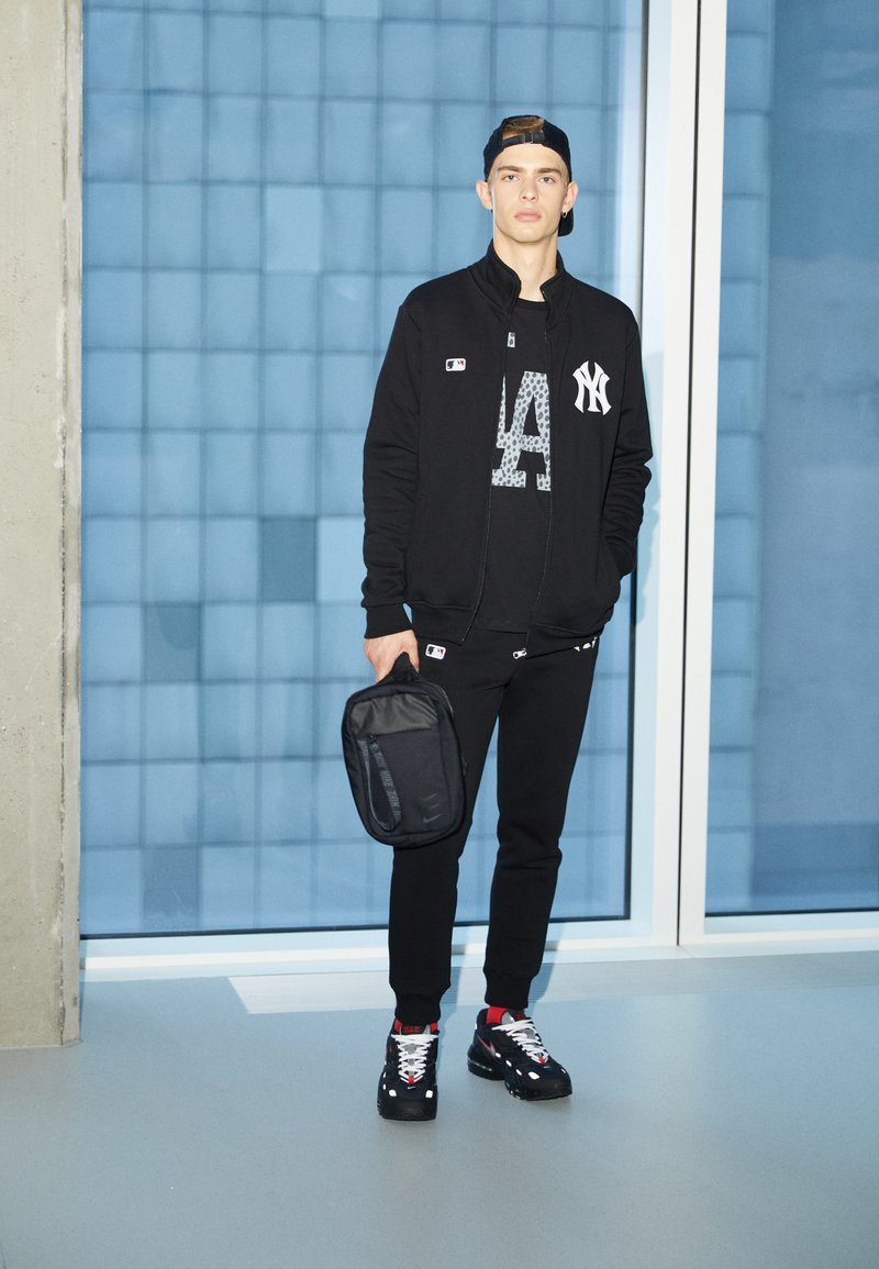 Black zip-up jacket with MLB logo, black graphic t-shirt, black joggers, and black sneakers with white accents. Holding a black backpack.