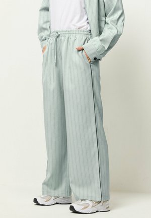 Personne portant un pantalon large à fines rayures vert clair avec taille élastique, veste assortie, chemise blanche et baskets blanches.