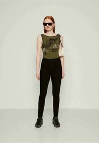 Γυναίκα με μακριά κόκκινα μαλλιά που φορά μαύρα γυαλιά ηλίου, ελαιώδης πράσινη χωρίς μανίκια μπλούζα, μαύρο παντελόνι, αθλητικά παπούτσια και κρατώντας μια μικρή λευκή τσάντα ώμου.