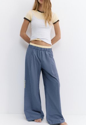 Nő áll egy fehér fal előtt, bézs-fekete rövid ujjú pólót visel világosszürke ujjatlan felső alatt, és laza kék kockás nadrágot rugalmas derékkal.