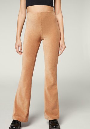 Frau trägt hoch taillierte, camel-farbene Samt-Schlaghosen mit schwarzen Schuhen, stehend vor einem hellgrauen Hintergrund.