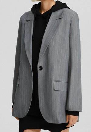 Femme portant un blazer gris à fines rayures sur un sweat à capuche noir et une jupe noire, debout devant un fond uni.