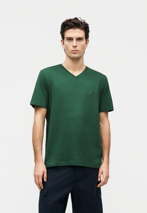 Fiatal férfi áll, sötétzöld V-nyakú pólót visel, melyen kis logó van a mellkasán, és sötét cargo nadrágot, sima háttér előtt.