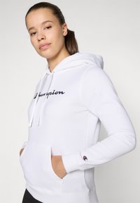 Hoodie blanc en mélange de coton avec poche frontale, capuche à cordons et logo "Champion" brodé en noir sur la poitrine.