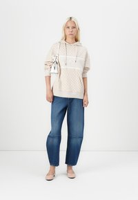 Beige Kapuzenpullover mit strukturiertem Muster, weißen Akzenten und Kängurutasche, kombiniert mit lässigen blauen Jeans und hellrosa Ballerinas.