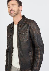 Braune Lederjacke mit strukturiertem Finish, ausgestattet mit einem Reißverschluss vorne, zwei Brusttaschen und gesteppten Schulterakzenten.