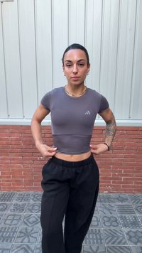 Egy tetovált karú, alakformált nő a szürke atlétikai crop topját igazítja, miközben mintás csempéken áll egy téglafal és fémfal előtt.