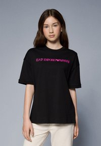 Camiseta de algodón negra de manga corta, con un logo en rosa "EA7 EMPORIO ARMANI" en el pecho. Ajuste simple y relajado.