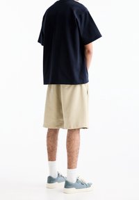 Homme en shorts beiges, t-shirt bleu marine, chaussettes blanches et baskets bleu clair se tenant devant un fond blanc uni, les mains dans les poches.