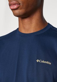 Tumši zila, apaļā kakla T-kreklu izgatavots no kokvilnas, ar zelta Columbia logo kreisajā krūtīs. Gluda tekstūra, klasiskā piegriezuma.