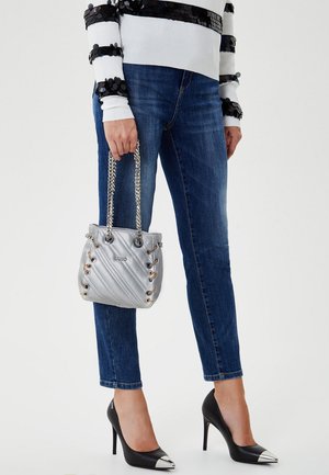 Mujer vistiendo jeans ajustados azules, tacones altos negros con puntas plateadas, suéter blanco con lentejuelas negras, sosteniendo un pequeño bolso acolchado plateado.