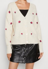 Krämfärgad cardigan med rosa rosbroderi, v-ringad och med knapplåsning. Bärs över en svart topp och matchas med en svart lädrokjol.