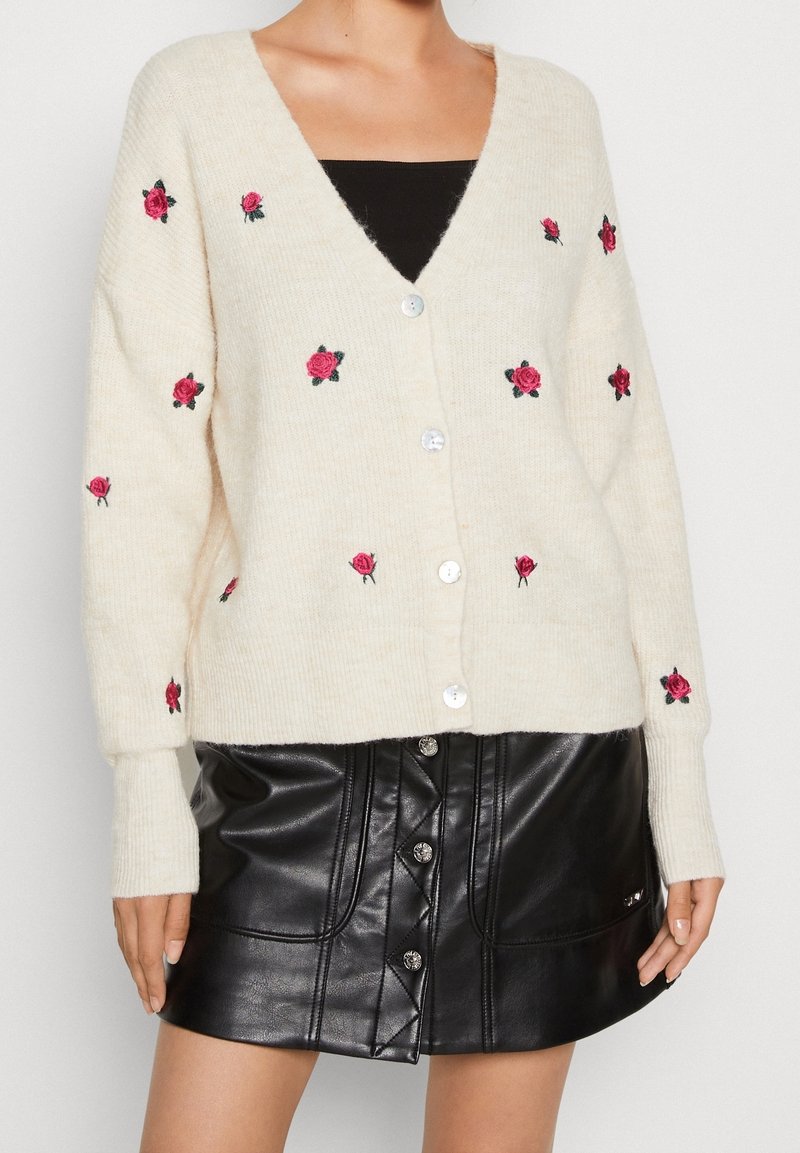 Krämfärgad cardigan med rosa rosbroderi, v-ringad och med knapplåsning. Bärs över en svart topp och matchas med en svart lädrokjol.