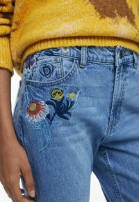 Helle blaue Jeans mit floralem Stickerei in Rot, Gelb und Blau auf der rechten Hüfte; verfügt über einen Metallknopf und ein klassisches Fünf-Taschen-Design.