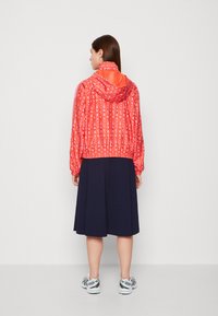 Giacca con cappuccio a motivo rosso, con polsini elasticizzati, di design cropped, abbinata a una gonna blu scuro e sneakers bianche, mostrata di spalle.