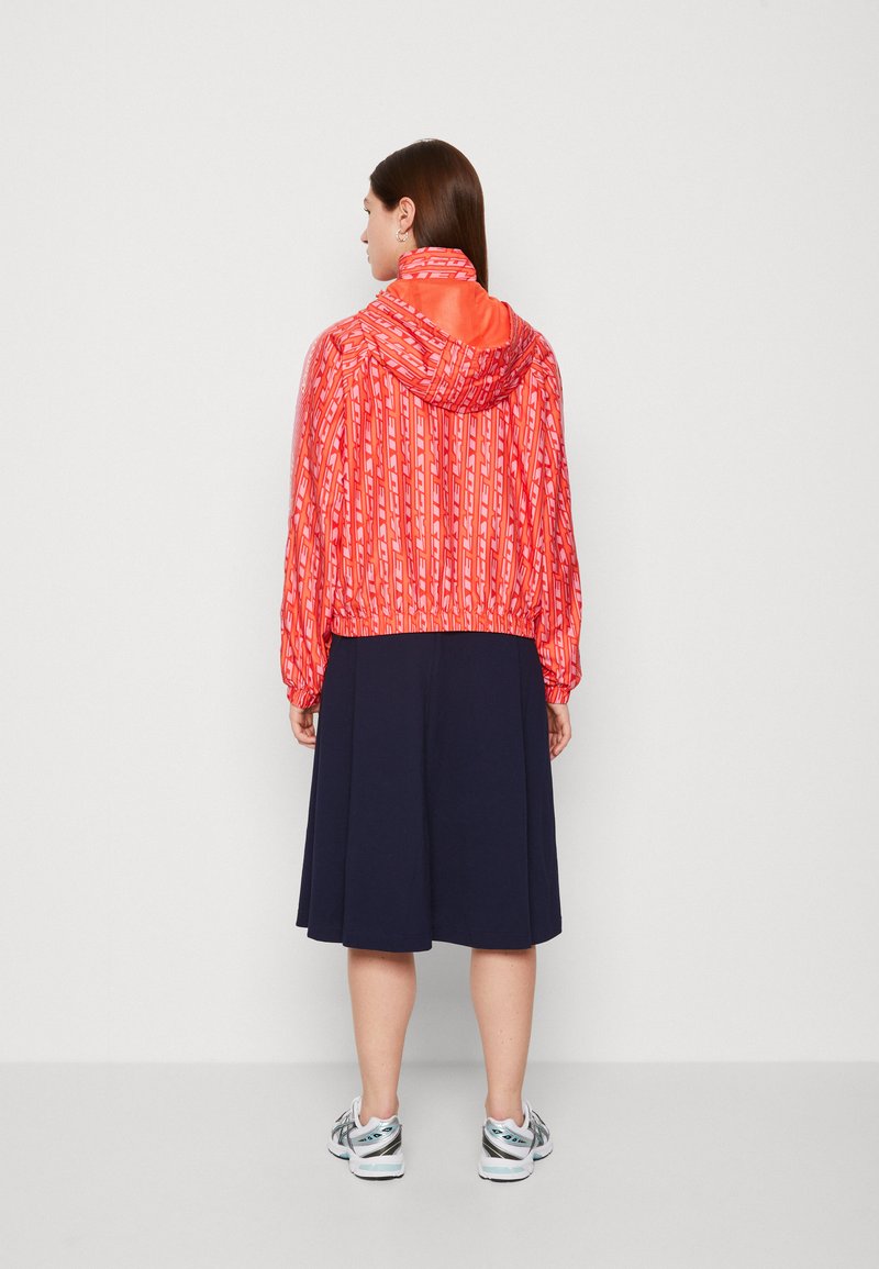 Chaqueta con capucha de patrón rojo y puños elásticos, diseño cortado, combinada con una falda azul marino y zapatillas blancas, mostrada desde la parte de atrás.