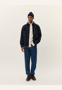 Marine-stripete skjortejakke i teksturert stoff, lagdelt over en beige skjorte, kombinert med blå jeans og mørkebrune sko, toppet med en marineblå lue.