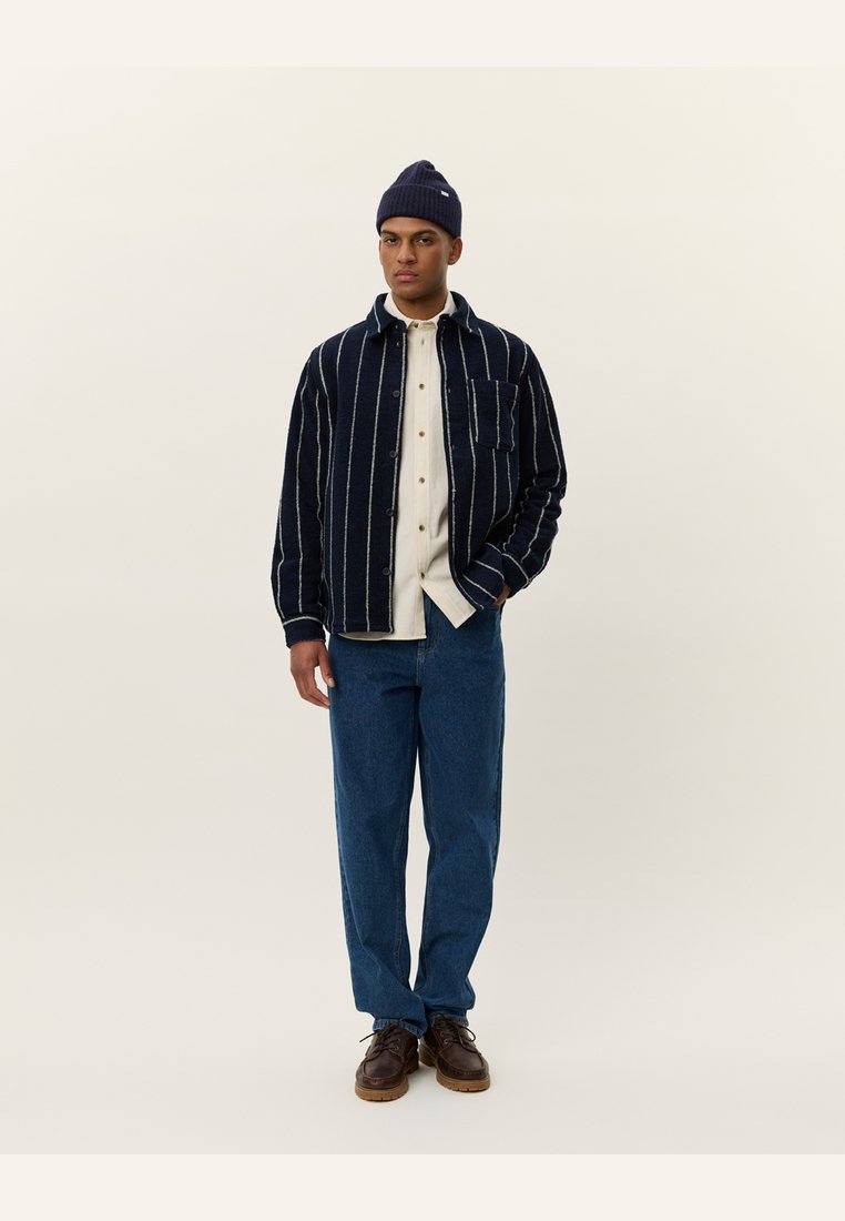 Marine-stripete skjortejakke i teksturert stoff, lagdelt over en beige skjorte, kombinert med blå jeans og mørkebrune sko, toppet med en marineblå lue.