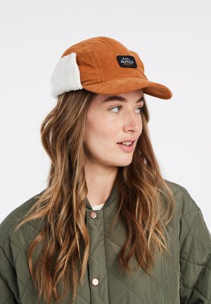 Bruine corduroy pet met witte faux fur oordoppen. Bevat een zwart geborduurd label met de tekst "PROTEST." De klep is licht gebogen.