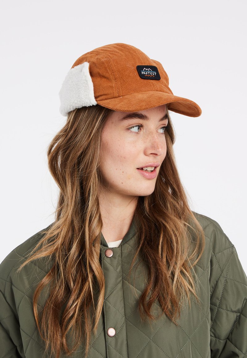 Bruine corduroy pet met witte faux fur oordoppen. Bevat een zwart geborduurd label met de tekst "PROTEST." De klep is licht gebogen.
