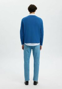 Personne de dos, portant un pull bleu, une chemise rayée, un jean bleu clair et des chaussures noires sur un fond blanc uni.