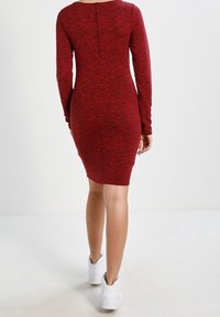 Robe bodycon rouge à manches longues avec un tricot texturé, présentant une fermeture à zip dans le dos et une silhouette ajustée, associée à des baskets blanches.
