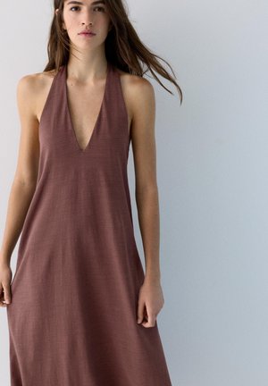 Junge Frau trägt ein ärmelloses, locker sitzendes braunes Kleid mit tiefem V-Ausschnitt und steht vor einem schlichten hellgrauen Hintergrund.