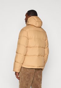 Bež puffer jakna s kapuljačom, koja ima kvadratne dijelove i glatku teksturu. Nosena je preko smešnih žamijskih hlača.