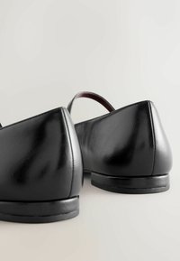 Pair of black leather flat shoes with rounded heels shown from the back on a plain light background.