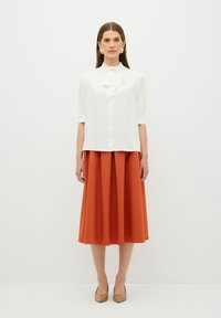 Blusa bianca con scollo a fiocco e maniche corte a pieghe, abbinata a una gonna arancione lunga fino alla caviglia. Tacchi beige completano l'outfit.
