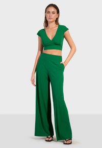 Zeleni kratki top s kap sleeves i dubokim V-izrezom, uparen s hlačama visokog struka širokih nogavica koje imaju bočne džepove i glatku teksturu.