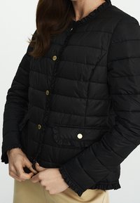 Černá prošívaná bunda s volány na okrajích, zlatými patentkami a předními kapsami. Tkanina má hladký povrch s lehkým vzhledem.