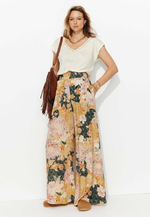 Pantalons larges à motifs floraux dans des teintes pastel avec une taille smockée, associés à un haut beige clair en V et un sac en cuir suédé brun en bandoulière.