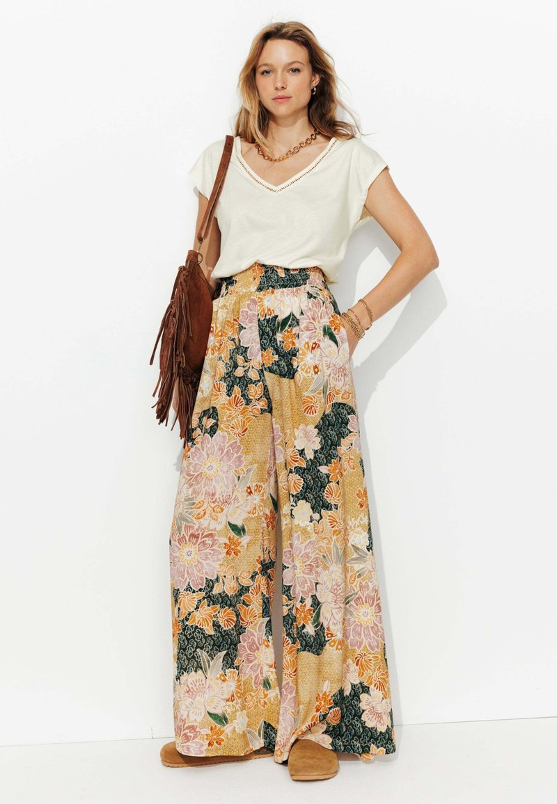 Pantalons larges à motifs floraux dans des teintes pastel avec une taille smockée, associés à un haut beige clair en V et un sac en cuir suédé brun en bandoulière.