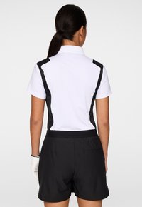 Vrouw gezien van achteren, gekleed in een witte polo met korte mouwen en zwarte zijpanelen, zwarte shorts en een witte handschoen om haar linkerhand.