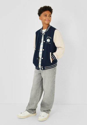 Jongen draagt een marineblauw en crème varsityjack, gestreept shirt, losse lichtgrijze spijkerbroek en witte sneakers, staand met handen in zakken.