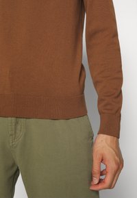 Homme portant un pull à manches longues marron et un pantalon vert olive, la main droite détendue sur le côté, sur un fond neutre.