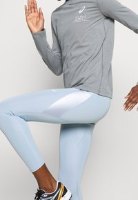 Hellgraues langärmeliges Trainingsoberteil mit grafischem Text auf der Vorderseite; hellblaue Leggings mit glänzendem Einsatz und figurbetontem Design.