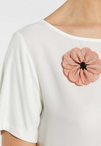 Hvid kortærmet top med en tekstureret, lyserød stofblomst-nål med sorte perleaccentuer, placeret på venstre skulder.