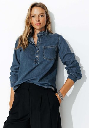 Chemise en denim bleu moyen avec une patte de boutonnage, un col et une poche poitrine ; manches longues avec des poignets retroussés. Associée à un pantalon noir.